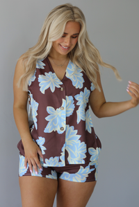 Woman wearing a brown, blue, and off white floral two piece set featuring a vest style top with functional button closures in the front and shorts. The photo is taken in front of a white wall.
