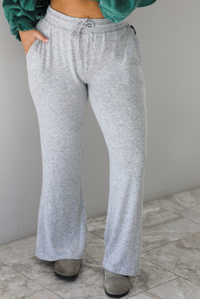 Woman model wearing heather grey lounge pants that have a high, elastic and drawstring waistline with functional pockets on either side, and a flowy hem. The photo focuses on the pants. The pants are paired with a darker grey slide on shoe. The photo is taken indoors against a white wall with white marble flooring. 