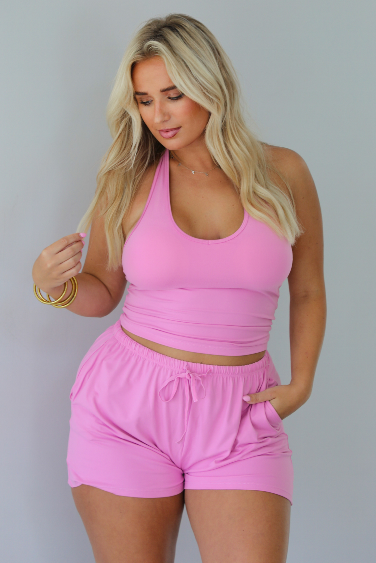 A woman wearing a matching pink activewear set consisting of a halter crop top and drawstring shorts, standing indoors against a white wall.