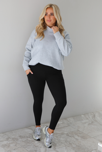 Woman wearing a grey turtleneck sweater tucked into high waisted black leggings. The outfit is paired with silver sneakers. The photo is taken indoors against a white wall with white marble flooring.