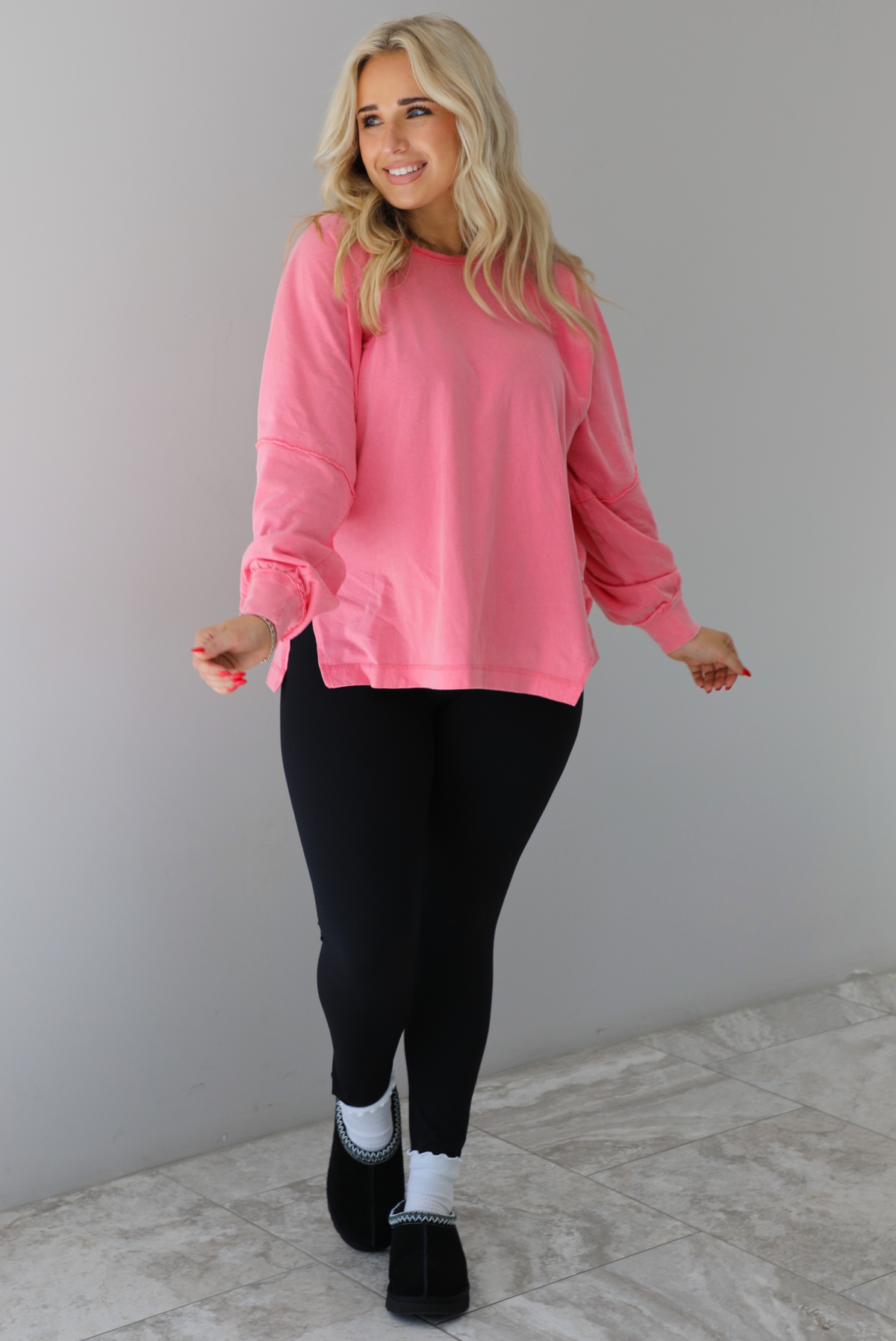 Woman wearing a pink oversized long sleeved top that has a high neckline, exposed seam details, split hem details on either side, and is lightweight. The top is modeled with a pair of fitted black leggings with black uggs and socks. The photo is modeled indoors against a white wall with white marbled flooring. 