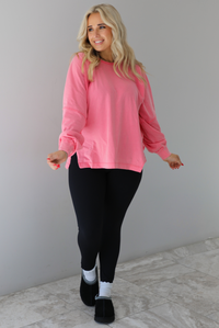 Woman wearing a pink oversized long sleeved top that has a high neckline, exposed seam details, split hem details on either side, and is lightweight. The top is modeled with a pair of fitted black leggings with black uggs and socks. The photo is modeled indoors against a white wall with white marbled flooring. 
