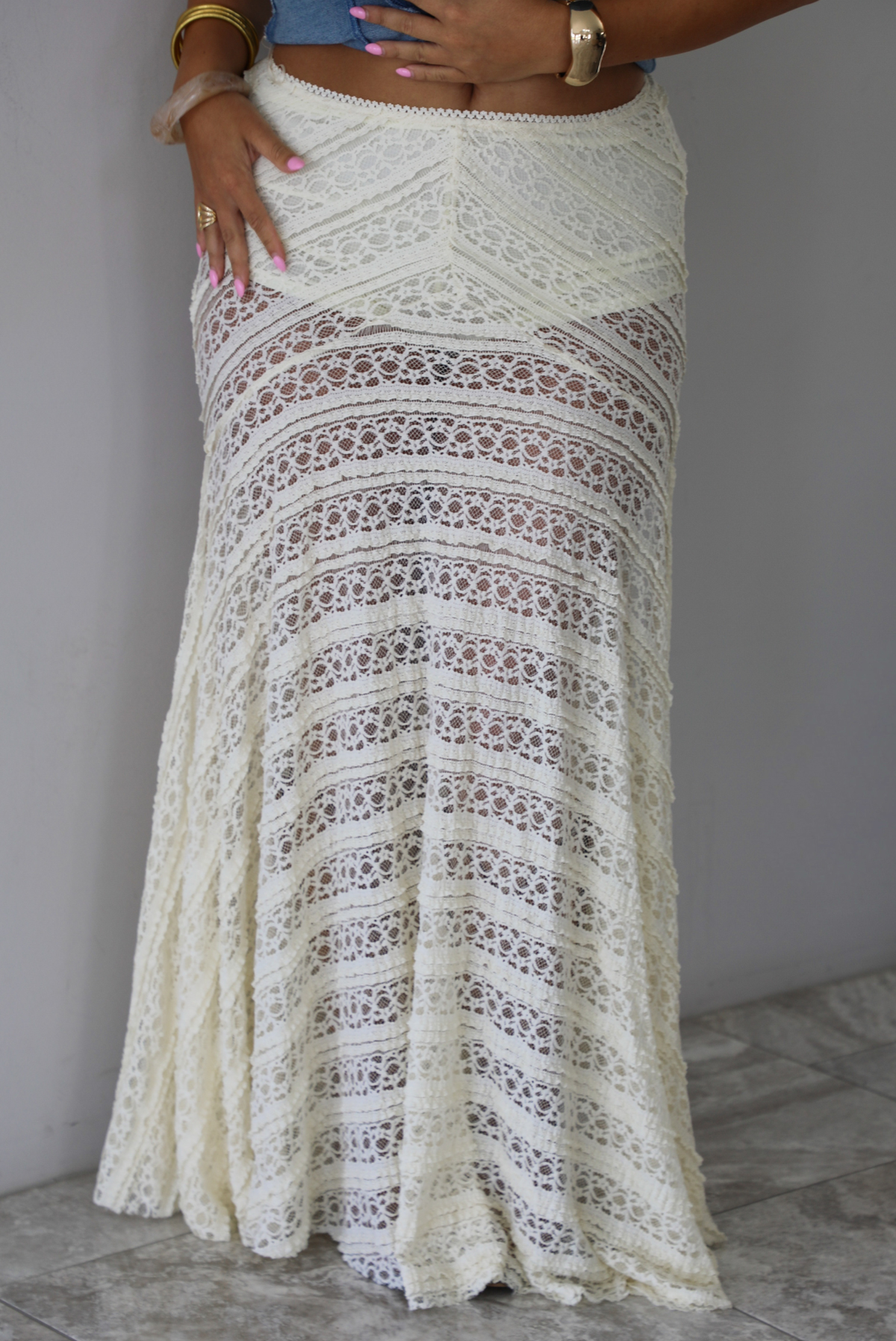 An off-white crochet lace maxi skirt with a tiered design, shown on a model indoors against a white wall with white marble flooring. 