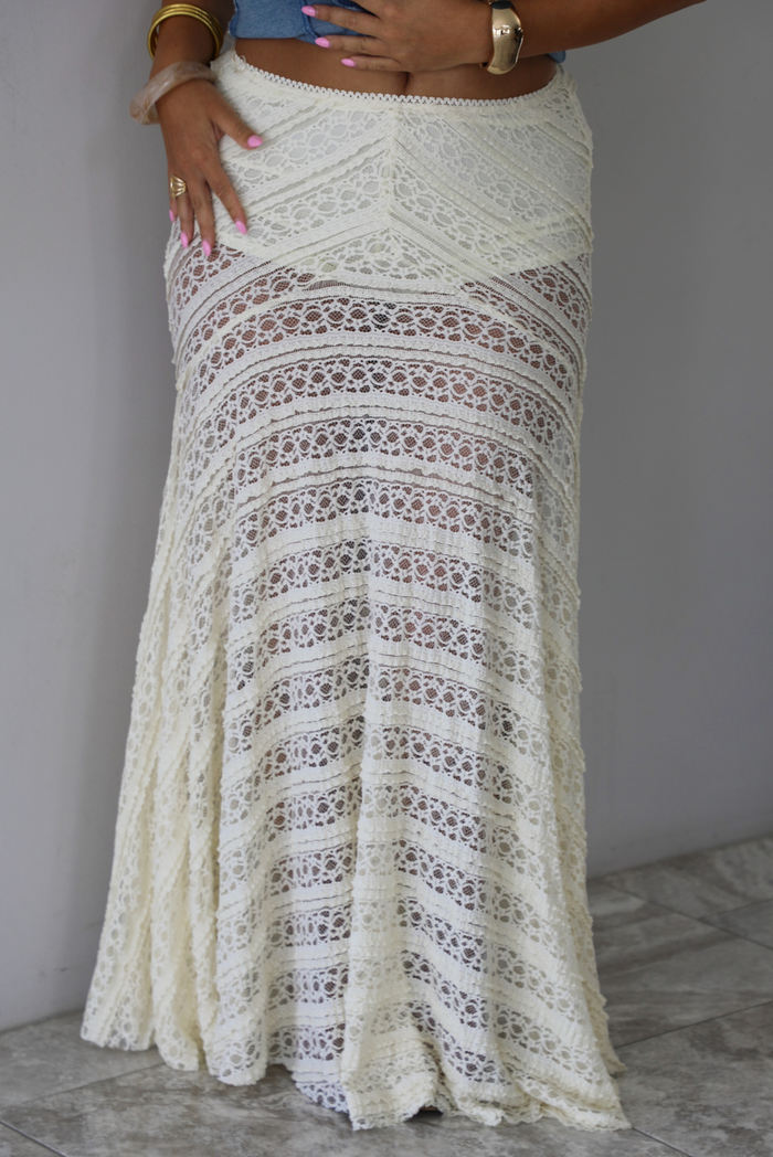 An off-white crochet lace maxi skirt with a tiered design, shown on a model indoors against a white wall with white marble flooring. 