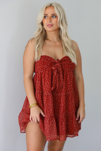 A woman wearing a rust mini length dress with a cream floral and leaf tiny print design throughout, has a strapless design with a tie detail in the center of the bust, with a baby-doll dress cut that's fitted at the bust with a flowy skirt, standing indoors against a white wall.