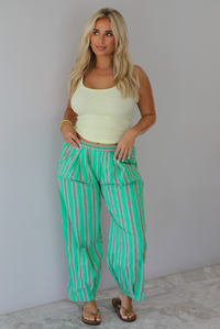A woman wearing a pale yellow tank and mid-rise pants that have a green, brown, lime green, and tan striped pattern, which have an elastic waistline, functional pockets on either side, and cinched details at the ankles, standing indoors against a white wall with white marble flooring. 
