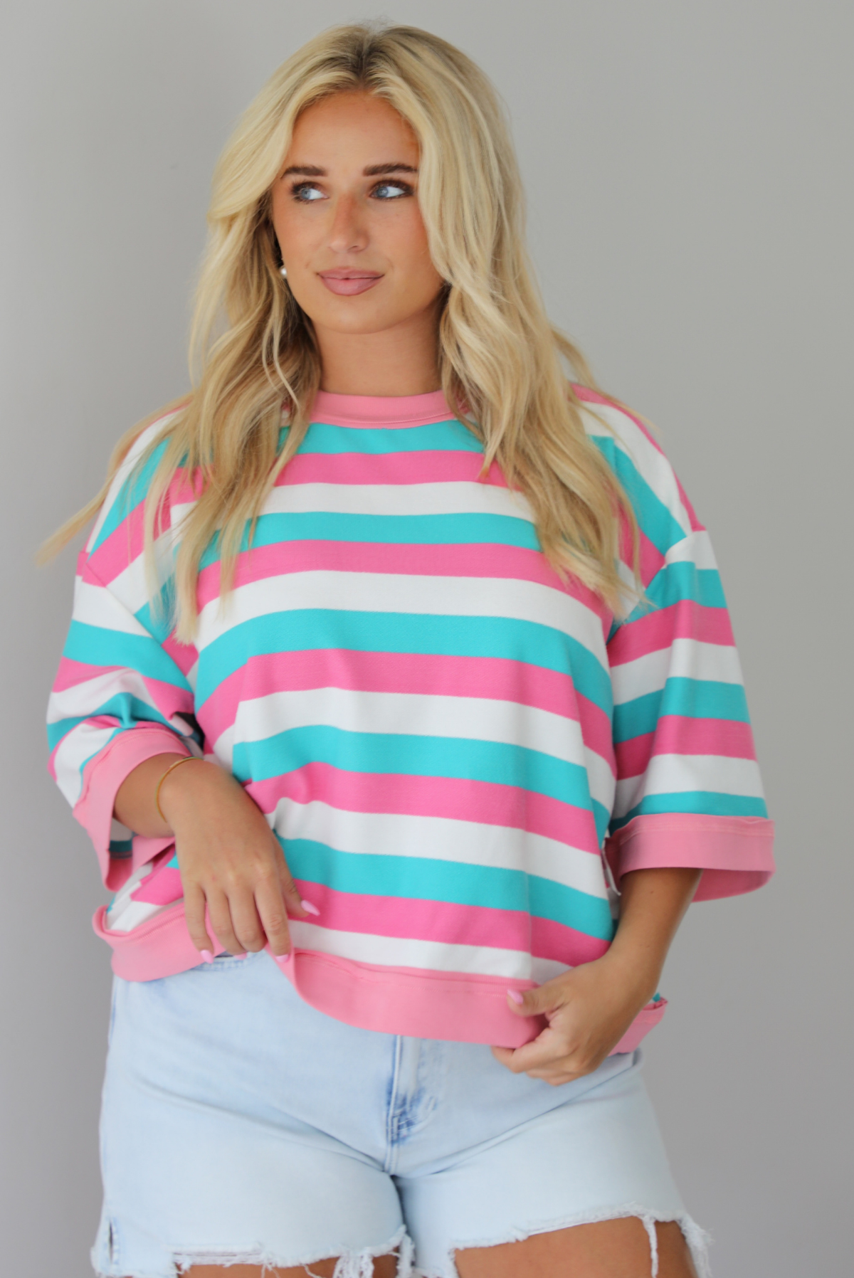 Woman wearing turquoise, pink, and white striped top with light wash denim shorts indoors against a white wall. The photo shows the top untucked to show the full length. 