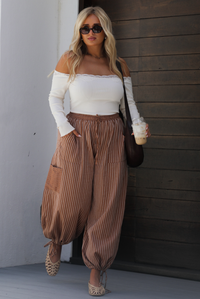 A woman wearing a cream off the shoulder long sleeved top tucked into a pair of camel and cream striped pants, a brown shoulder bag, cream colored wicker flats, standing outdoors against a dark wooden door and white wall. 