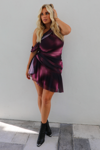 A woman modeling a short, one-shoulder mini dress in a deep purple with light purple design, paired with black ankle boots, standing outdoors against a white wall. 