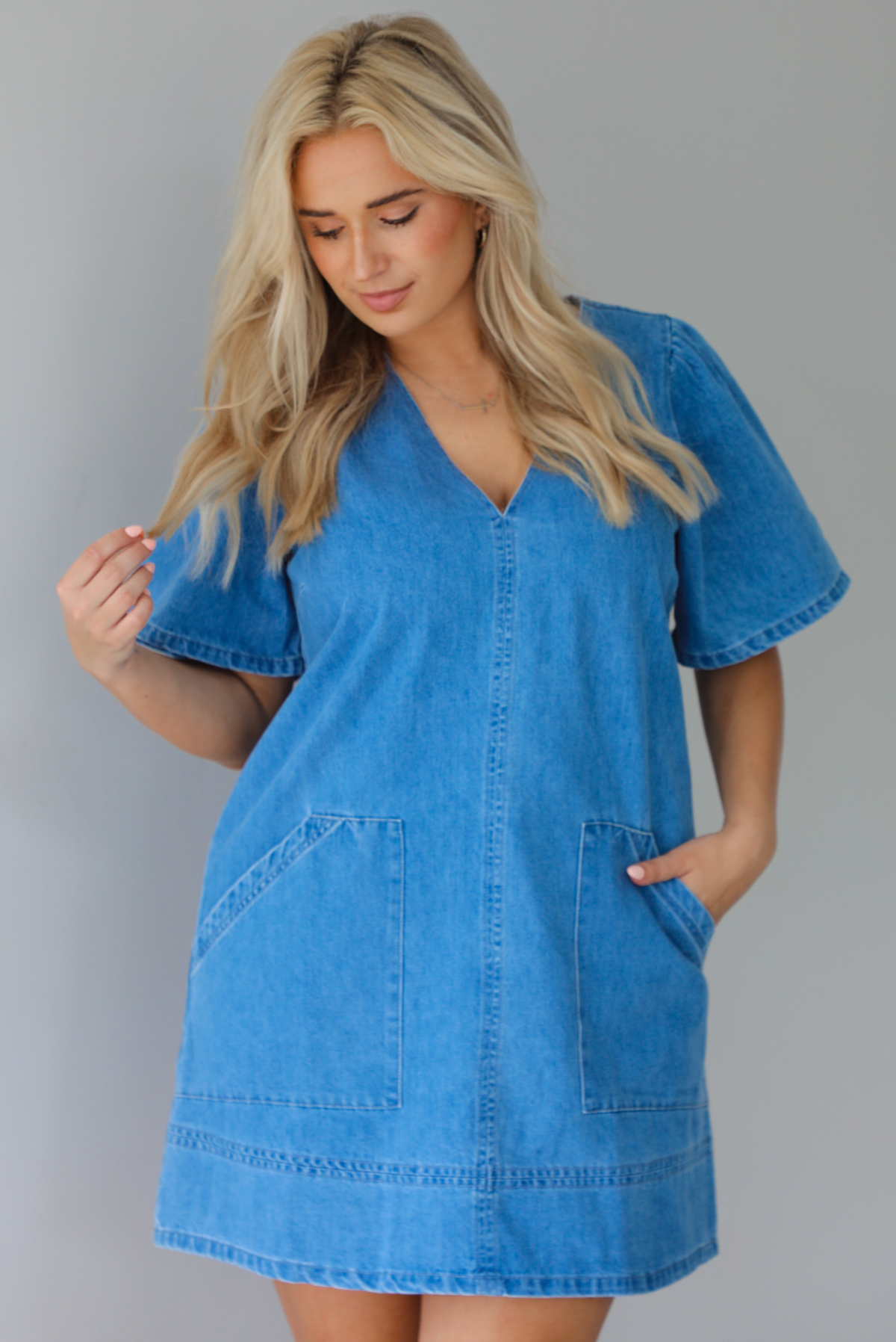 Woman wearing a v-neck, short sleeved, denim mini dress with functional pockets on either side. The photo is taken indoors against a white wall.