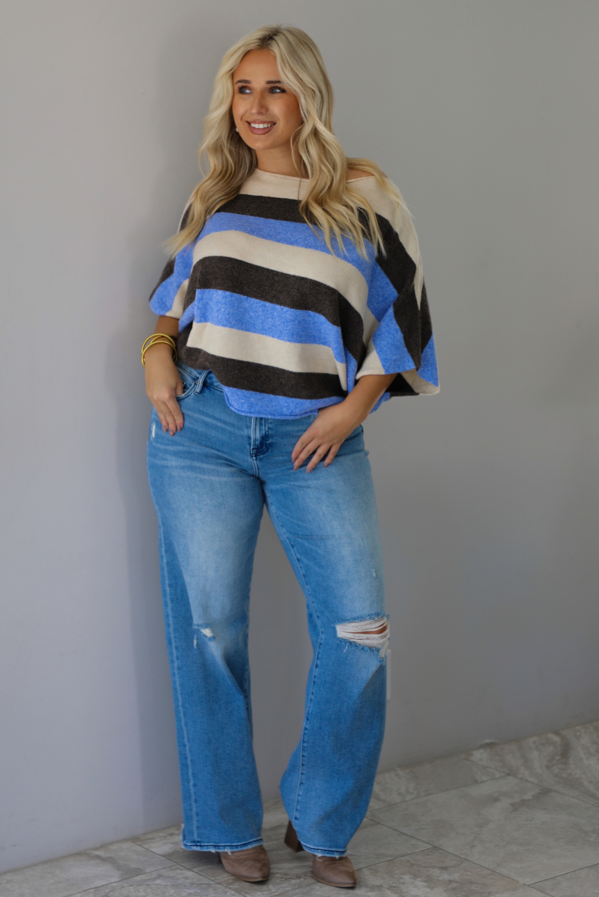 A woman wearing a cream, brown,, and blue striped knit top that has a boat-neck design, and quarter length flowy sleeves, paired with a pair of medium wash denim jeans, standing indoors against a white wall with white marble flooring. 