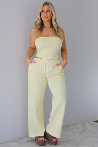 Woman wearing a pale yellow two piece set featuring a strapless tube top and high waisted, flared hem pants that have an elastic and drawstring waistline with functional pockets on either side. The photo is taken indoors against a white wall with white marble flooring. 