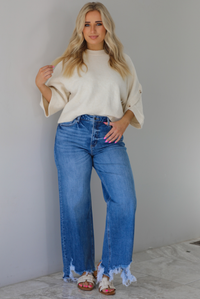 Woman wearing a cream colored quarter sleeve sweater that has ribbed details at the neckline, on the sleeves, and on the hem, with non-functional button closures on the sleeves. The sweater has a relaxed fit and is paired with a medium wash, high waisted, distressed hem denim with sand colored sandals. The photo is taken indoors against a white wall with white marble flooring. 