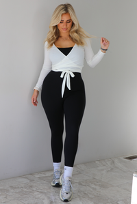 Woman wearing a white cropped wrap style fitted top over a black fitted jumpsuit. The outfit is paired with white ankle socks and silver sneakers. The photo is taken indoors against a white wall with white marble flooring. 