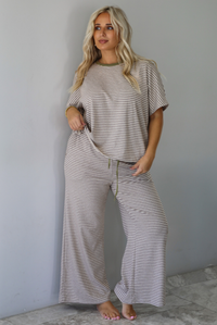 A woman modeling a matching two-piece loungewear set consisting of a short-sleeve top and wide-leg pants, both featuring thin beige and olive green horizontal stripes, with an olive green drawstring waist, standing indoors against a white wall with white marble flooring. 
