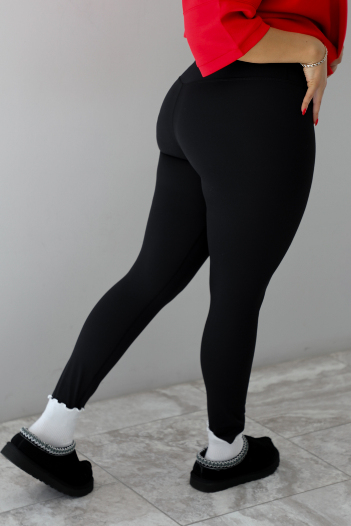 Woman model wearing a fitted pair of black leggings that are high waisted, soft, and super stretchy. She's wearing them with a pair of white ruffled socks and black slip on ugg boot shoes. The photo shoes the back of the leggings, which look like the front.