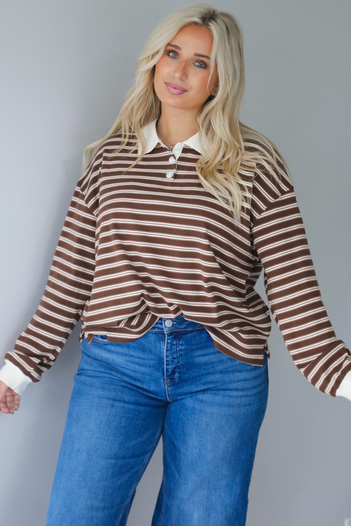A woman wearing a brown and cream striped collared quarter-button top, paired with dark, wide leg denim with a cuffed hem, standing indoors against a white wall.