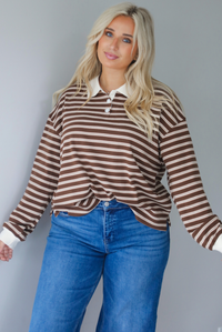 A woman wearing a brown and cream striped collared quarter-button top, paired with dark, wide leg denim with a cuffed hem, standing indoors against a white wall.