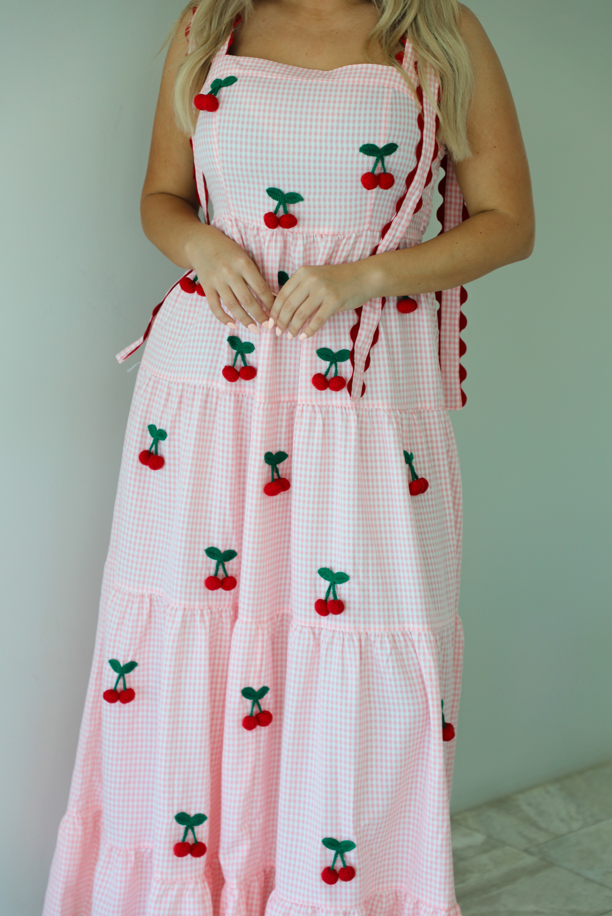 Woman wearing a pink and white gingham  maxi dress with thick straps, a fitted bodice, flowy skirt, and crocheted cherry details on the front indoors against a white wall with white marble flooring. 