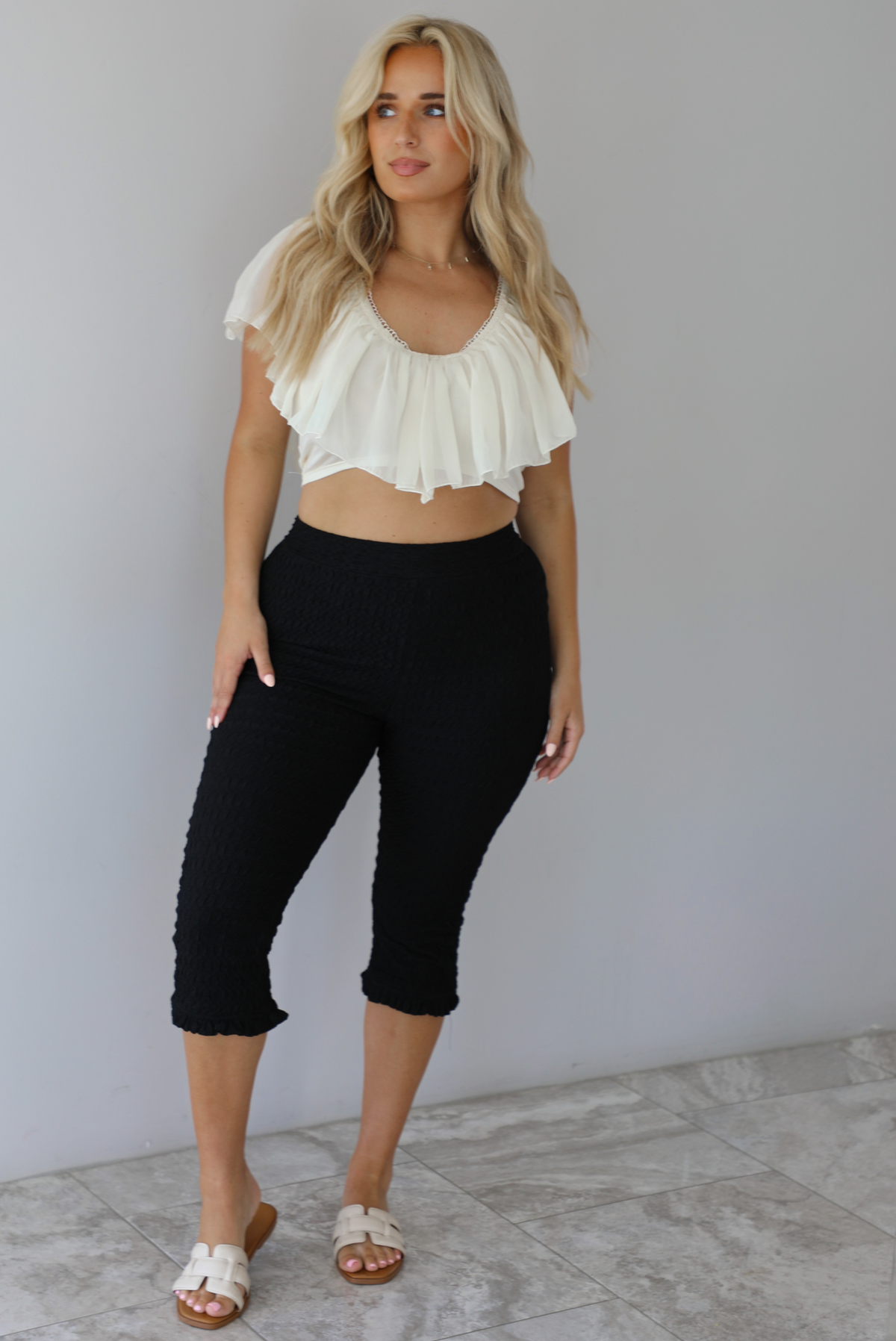 Woman wearing a cream ruffled cropped top with high waisted black, textured capri pants indoors against a white wall with white marble flooring. 