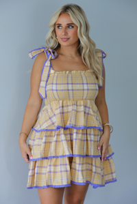 A woman modeling a yellow and purple plaid tiered mini dress with functional tie straps, standing indoors against a white wall.