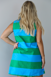 A woman wearing a sleeveless collared mini dress with bold horizontal stripes in green and blue, standing indoors against a white wall. The photo shows the back of the dress. 