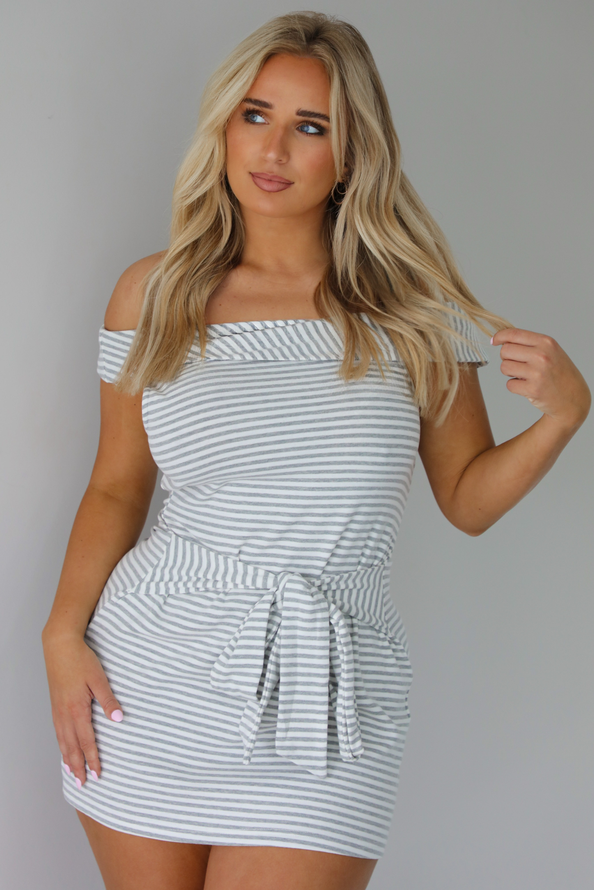 Woman wearing an off the shoulder grey and white striped mini dress that has a tie detail in the front. The photo is taken indoors against a white wall.