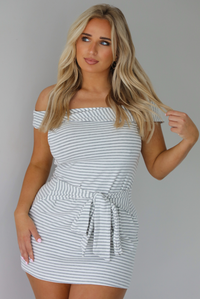 Woman wearing an off the shoulder grey and white striped mini dress that has a tie detail in the front. The photo is taken indoors against a white wall.
