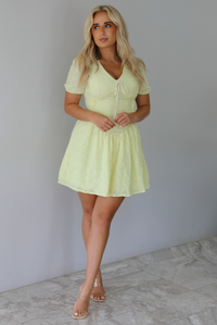 Woman wearing a light yellow mini dress with a floral eyelet design throughout, v-neckline, short sleeves, fitted drop waist bodice, and a flowy mini length skirt indoors against a white wall with white marble flooring. 
