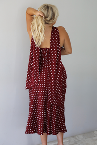 A woman shown from behind wearing a strapless maroon midi dress with a white polka dot pattern with a matching scarf, and has a zipper closure in the back, standing indoors against a white wall with white marble flooring. 