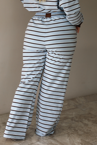 A woman shown from behind wearing a blue matching two piece set with dark brown and white striped details, featuring an oversized long sleeved top and high waisted loose fitting, flared leg pants, indoors against a white wall with white marble flooring. The photo shows a closeup of the back of the pants. 