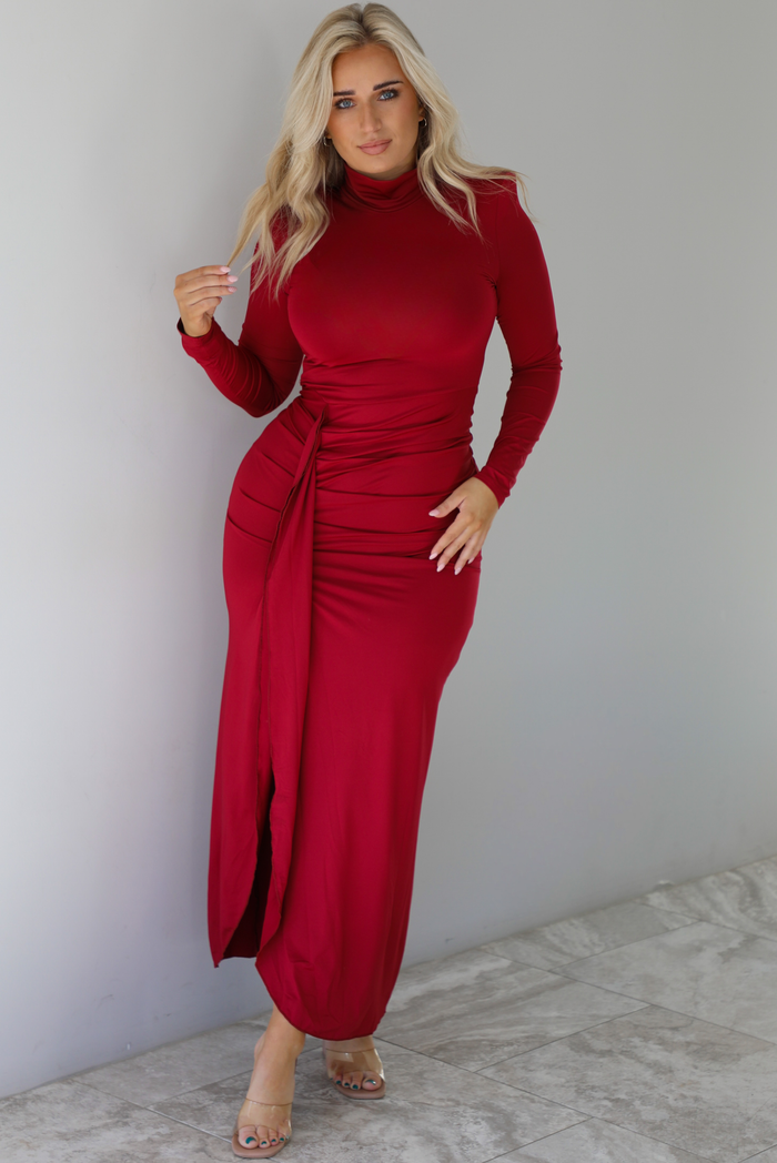 Woman wearing a red maxi dress that has a high neckline, shoulder pads, a fitted bodice, long sleeves, with a split hem detail on the left side. The photo is taken indoors against a white wall with white marble flooring. 