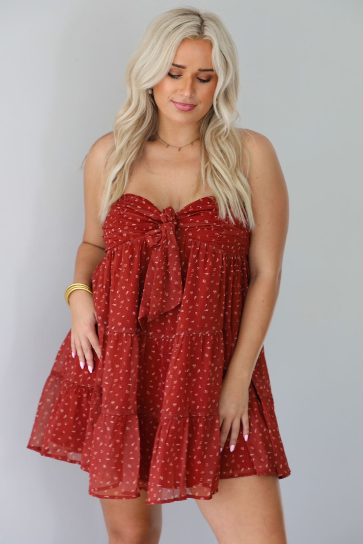 A woman wearing a rust mini length dress with a cream floral and leaf tiny print design throughout, has a strapless design with a tie detail in the center of the bust, with a baby-doll dress cut that's fitted at the bust with a flowy skirt, standing indoors against a white wall.