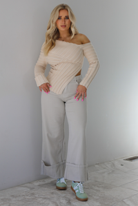 Woman wearing a cream colored ribbed textured knit top that has an asymmetrical hem with long sleeves in which one side is worn off the shoulder. The sweater is paired with sand colored high waisted, cuffed hem denim. The photo is taken indoors against a white wall with white marble flooring. 