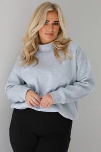 Woman wearing a grey turtleneck sweater tucked into high waisted black leggings. The photo is taken indoors against a white wall.