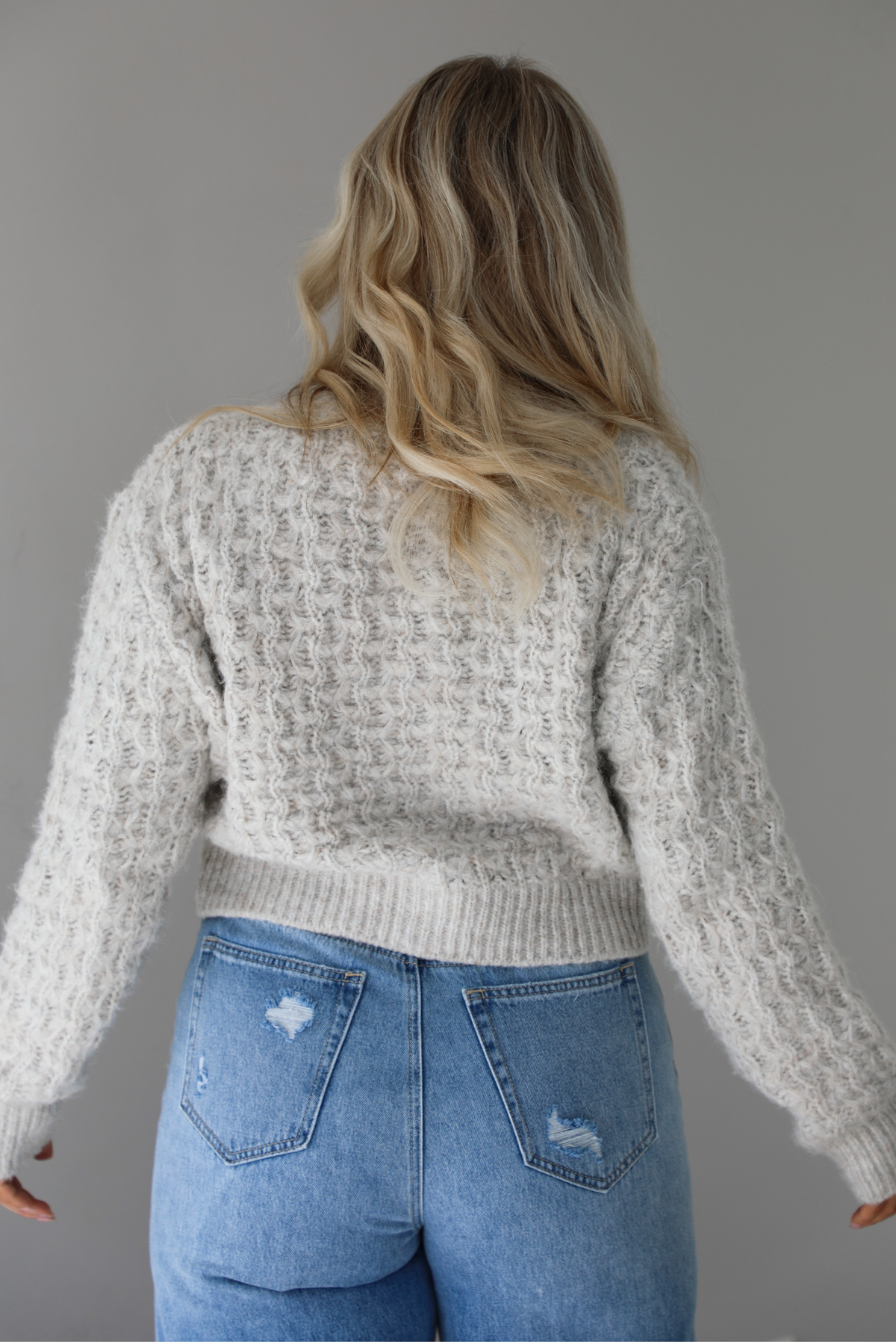 Woman wearing a light grey cardigan buttoned up with high waisted flared hem medium wash denim. The photo shows a closeup of the back of the cardigan and the texture of the cardigan, which resembles a loose textured cable knit. The photo is taken indoors against a white wall.
