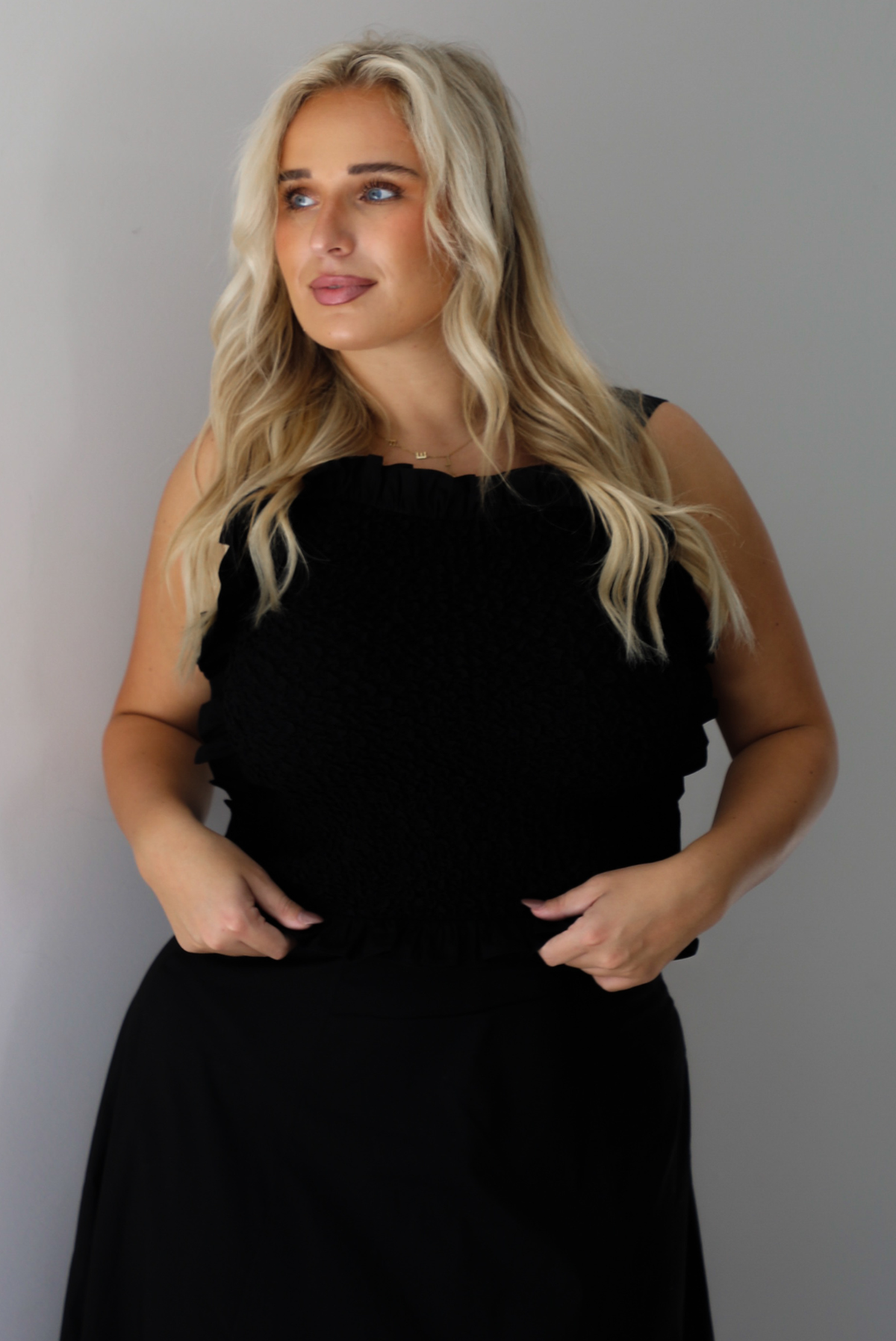 A woman wearing a two piece black set featuring a smocked detail top and high waisted maxi skirt, standing indoors against a white wall.