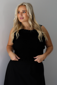 A woman wearing a two piece black set featuring a smocked detail top and high waisted maxi skirt, standing indoors against a white wall.