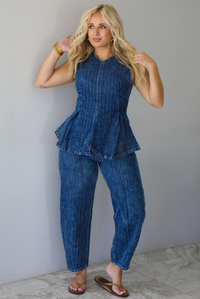A woman wearing a matching two piece dark wash denim set with a pin striped design on it featuring a sleeveless peplum style top and high waisted denim jeans, standing indoors against a white wall with white marble flooring. 