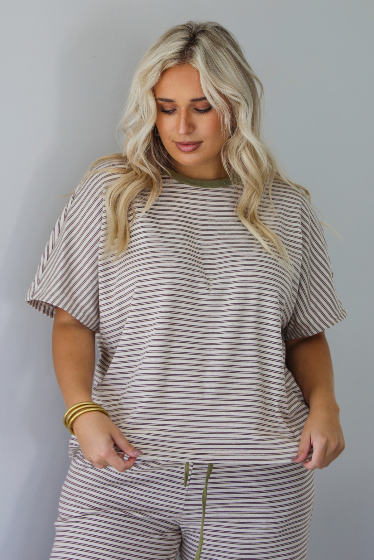 A woman wearing a matching short-sleeve striped lounge set in beige and olive green, featuring a crewneck top and drawstring pants, posed against a solid white wall. 