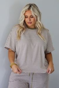 A woman wearing a matching short-sleeve striped lounge set in beige and olive green, featuring a crewneck top and drawstring pants, posed against a solid white wall. 