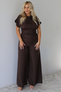 A woman modeling a dark brown short-sleeve jumpsuit with a high neckline and wide-leg pants, standing indoors against a white wall with white marble flooring. 