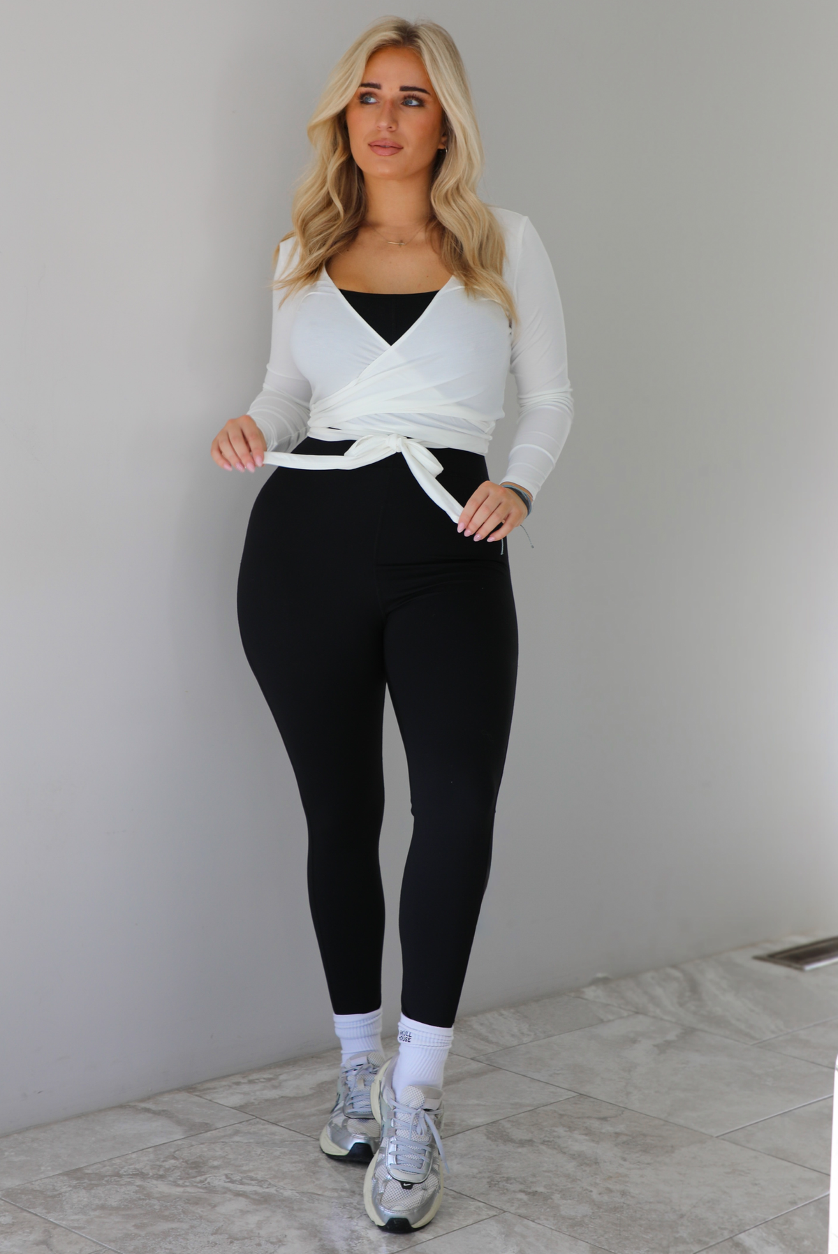 Woman wearing a white cropped wrap style fitted top over a black fitted jumpsuit. The outfit is paired with white ankle socks and silver sneakers. The photo is taken indoors against a white wall with white marble flooring. 