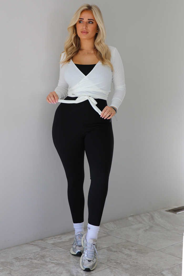 Woman wearing a white cropped wrap style fitted top over a black fitted jumpsuit. The outfit is paired with white ankle socks and silver sneakers. The photo is taken indoors against a white wall with white marble flooring. 