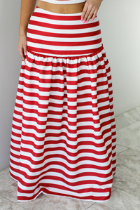 Woman wearing a red and white striped two piece set featuring a cropped top and maxi length, high waisted skirt. The photo is taken indoors against a white wall with white marble flooring, and shows a closeup of the front of the skirt. 