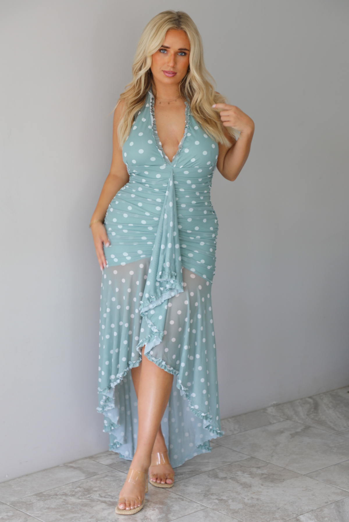 A woman modeling a light sage and white polka dot halter maxi dress with a high-low ruffled hem, standing indoors against a white wall with white marble flooring. 