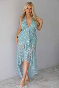A woman modeling a light sage and white polka dot halter maxi dress with a high-low ruffled hem, standing indoors against a white wall with white marble flooring. 