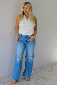 A woman modeling a cream sleeveless halter top and light-wash blue distressed flare jeans, standing indoors against a white wall with white marble flooring. 