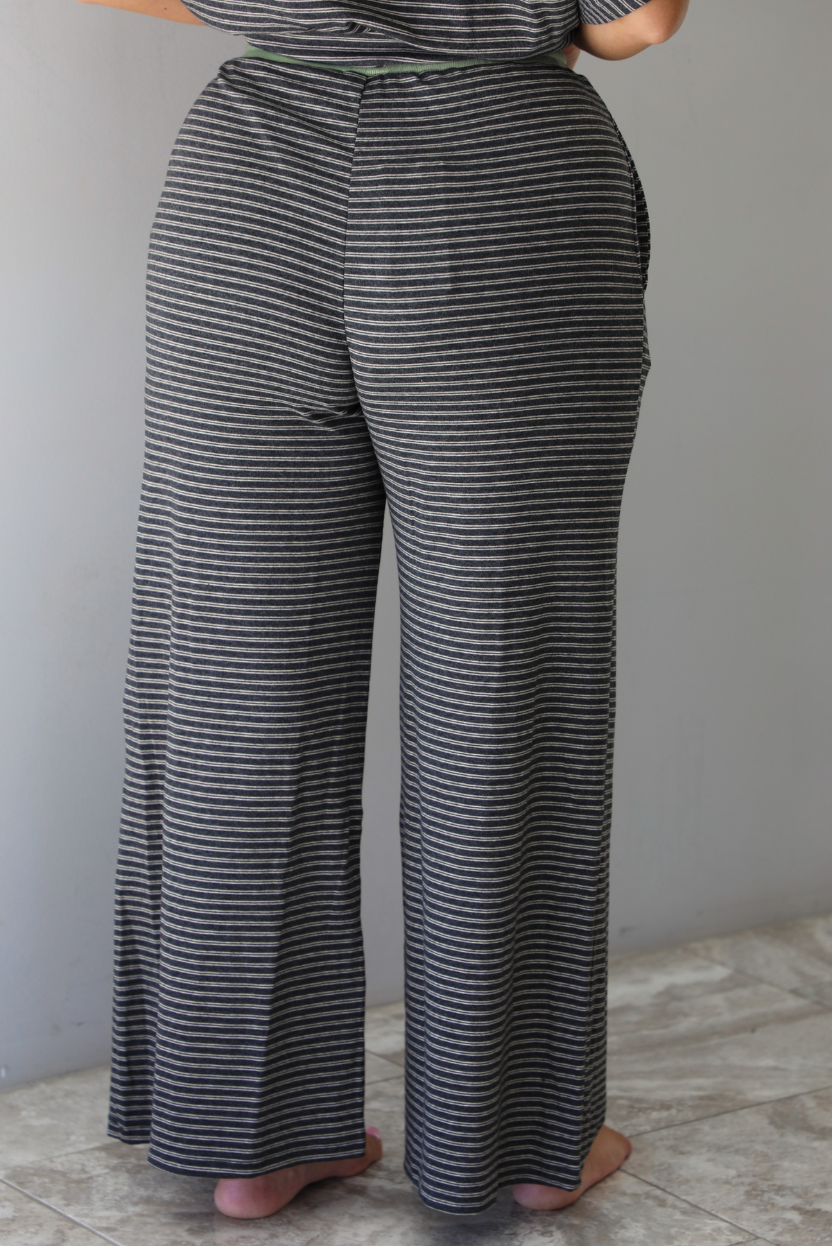 A woman shown from the waist down  and behind wearing a charcoal and white striped two piece matching set with green trim detailing at the neckline and waistline, featuring an oversized short sleeved top and high waisted pants, standing indoors against a white wall with white marble flooring. The photo shows a closeup of the back of the pants. 