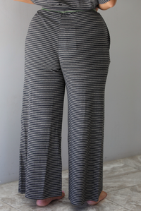 A woman shown from the waist down  and behind wearing a charcoal and white striped two piece matching set with green trim detailing at the neckline and waistline, featuring an oversized short sleeved top and high waisted pants, standing indoors against a white wall with white marble flooring. The photo shows a closeup of the back of the pants. 